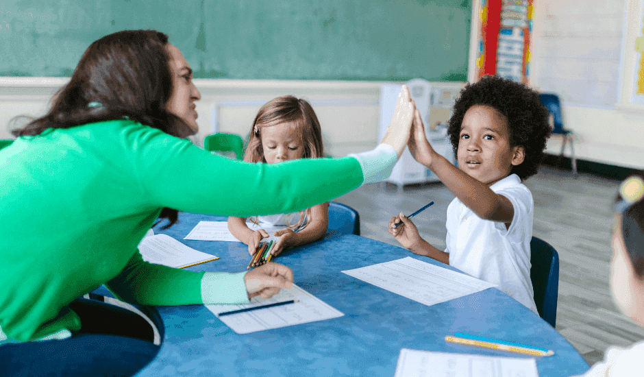 Teaching assistant sat at a table, helping young children with their learning.
