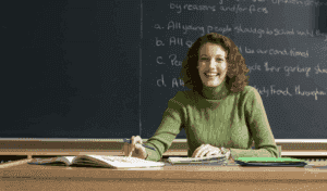 Dedicated Teacher Sitting At A Desk In Front Of A Chalkboard, Smiling And Holding A Pen, With Open Books And Papers On The Desk To Show There Are Great Teachers.