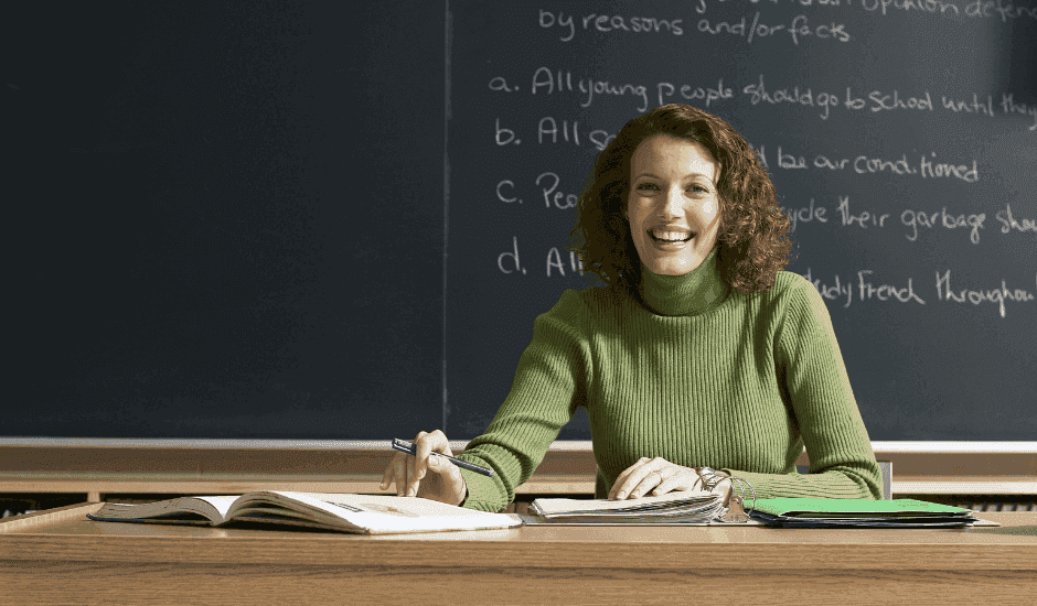 Dedicated teacher sitting at a desk in front of a chalkboard, smiling and holding a pen, with open books and papers on the desk to show there are great teachers.
